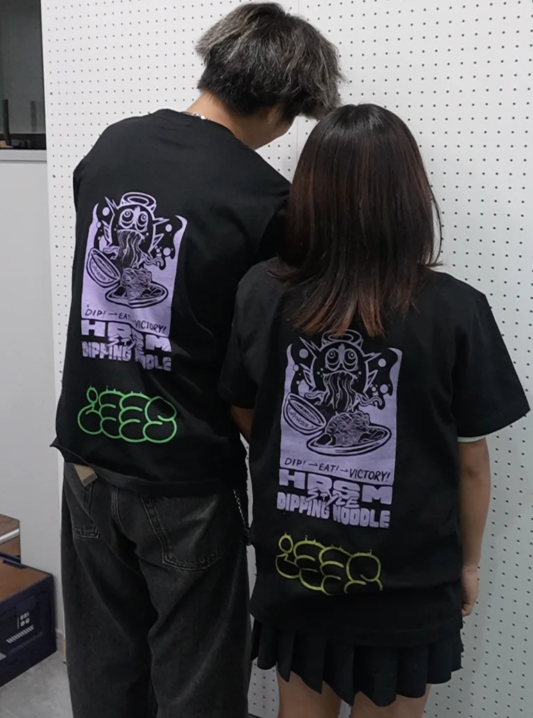 同じデザインで下部テキストだけ色違いのTシャツを着たカップルの後ろ姿 – EDITION SEE? 広島
